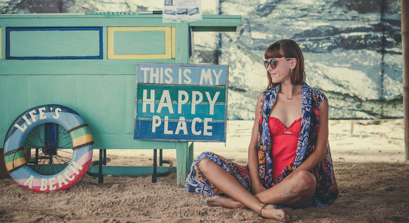 Woman sitting at beach smiling with a sign that says "This Is My Happy Place: